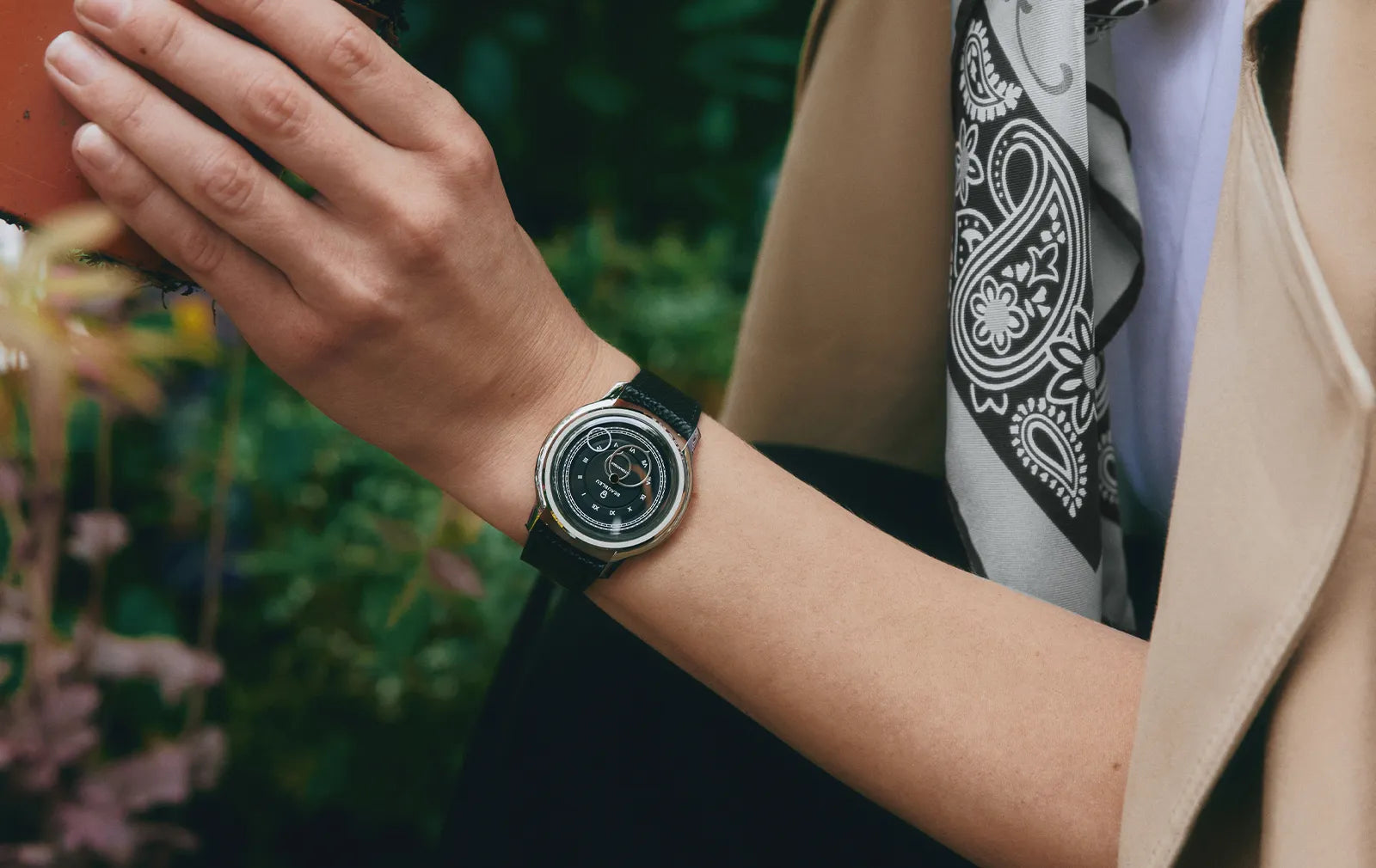Poignet portant une montre La Pièce Graphite et bracelet grainé noir, photographié dans un jardin fleuri
