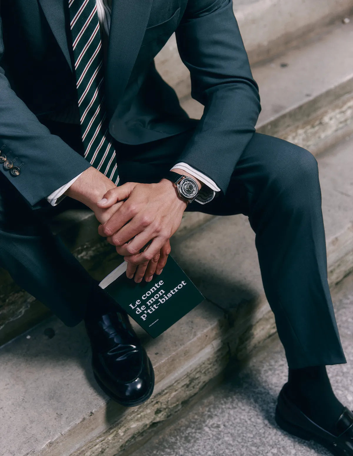 Homme assis sur des marches en costume sombre, portant une montre La Pièce à cadran noir et bracelet métallique