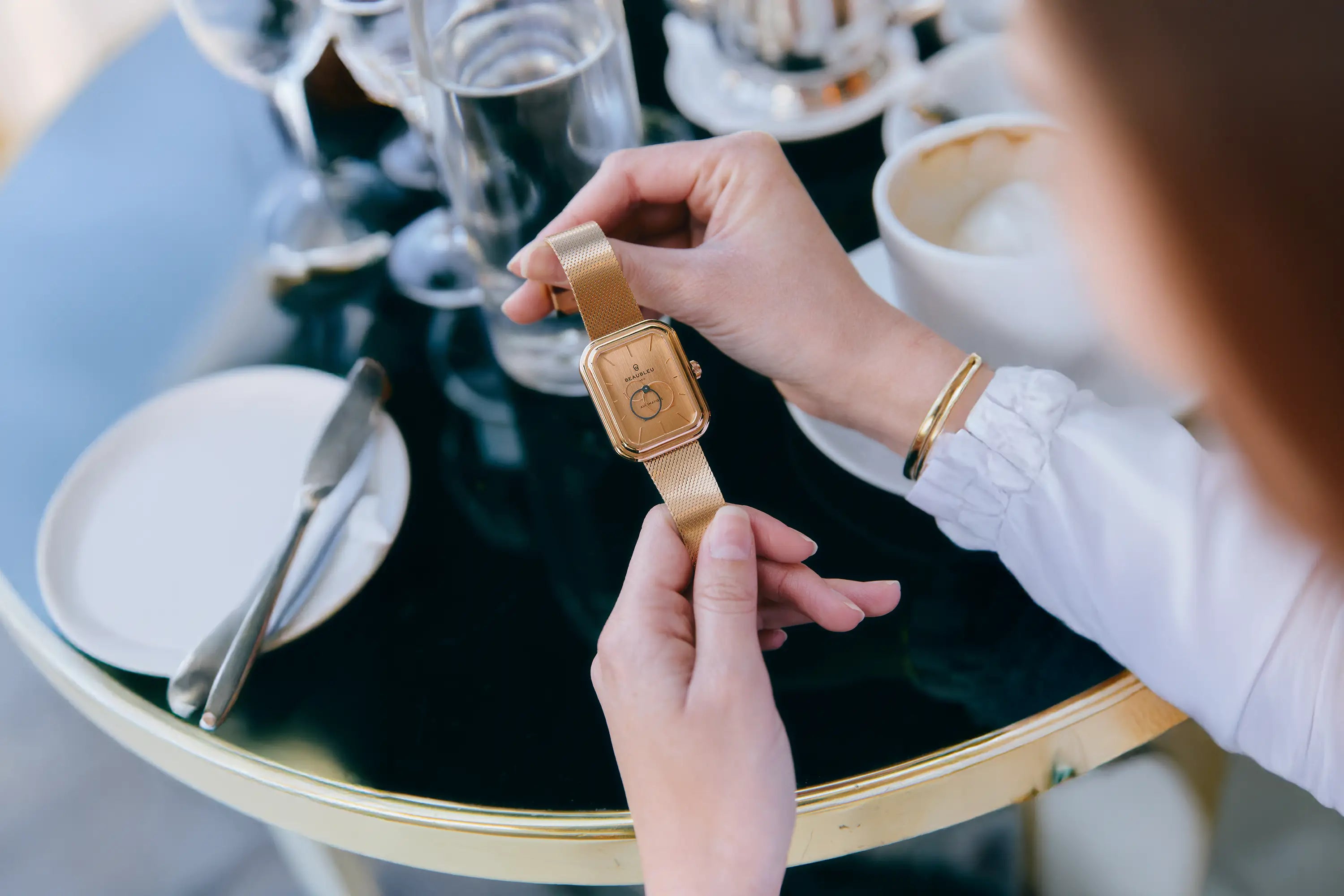 Femme qui tient entre ses mains une montre or rose avec un bracelet en acier or rose à la terrasse d'un café