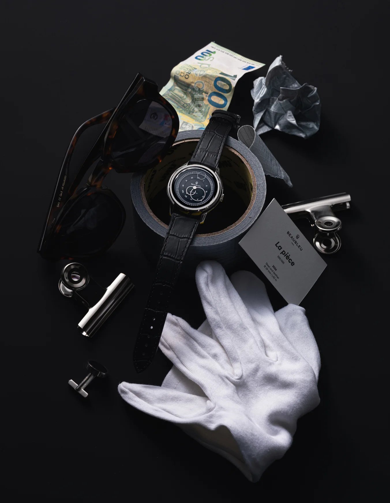 Montre La Pièce Graphite de Beaubleu cadran gris posée sur un ruban adhésif avec plusieurs accessoires autour