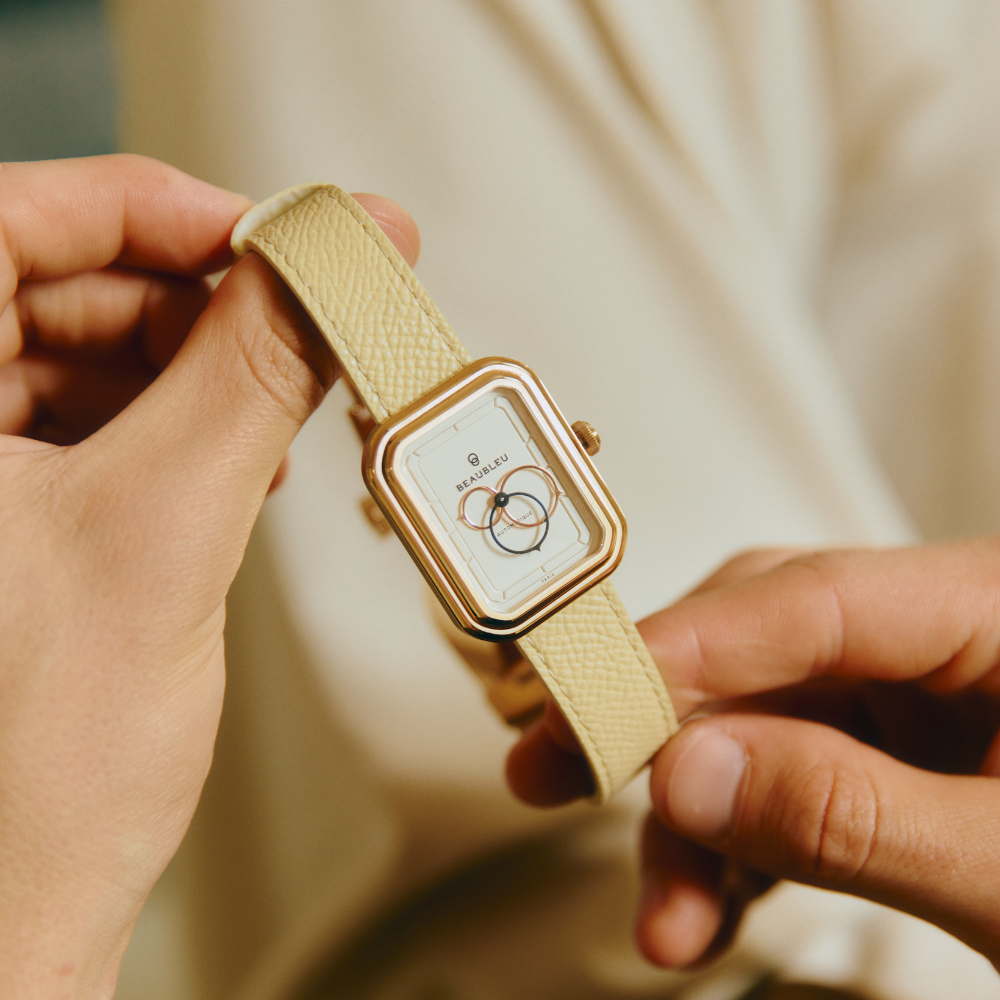 Montre carrée au cadran blanc, tenue entre deux doigts, avec un bracelet en cuir crème texturé