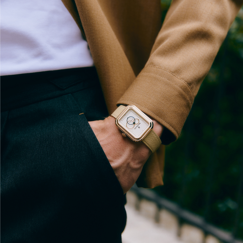 Montre carrée au boîtier doré et cadran blanc, portée sur un poignet masculin habillé d'une veste marron