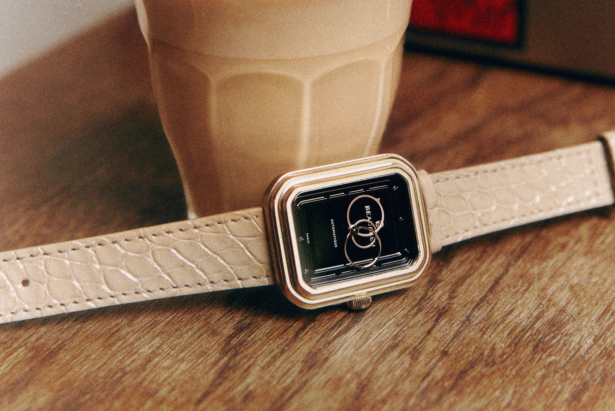 Gros plan sur une montre Beaubleu à cadran noir associée au bracelet Flat White en cuir d’alligator crème, près d’un café latte