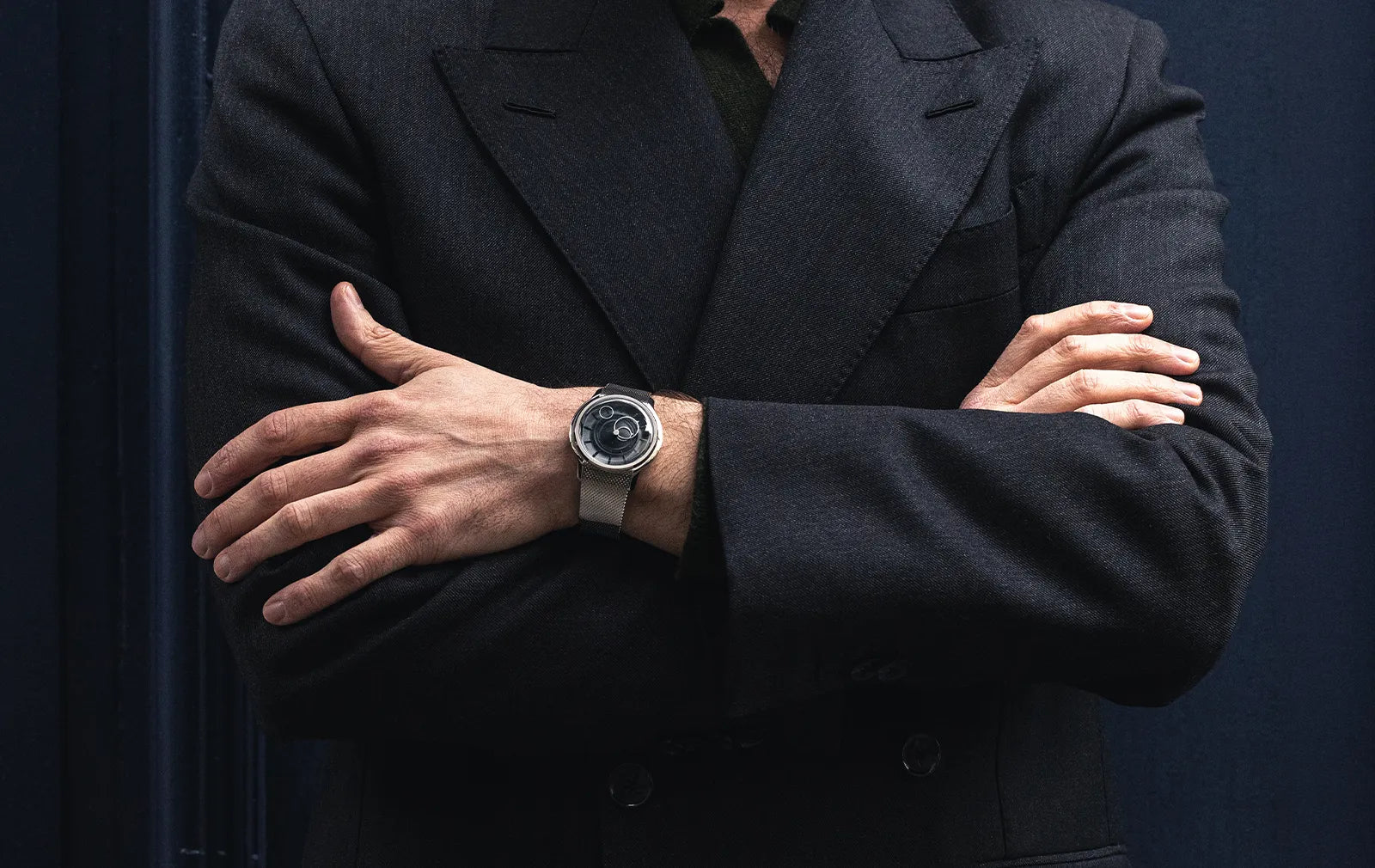Homme en costume noir croisant les bras, mettant en valeur une montre à cadran noir de la collection La Pièce de Beaubleu