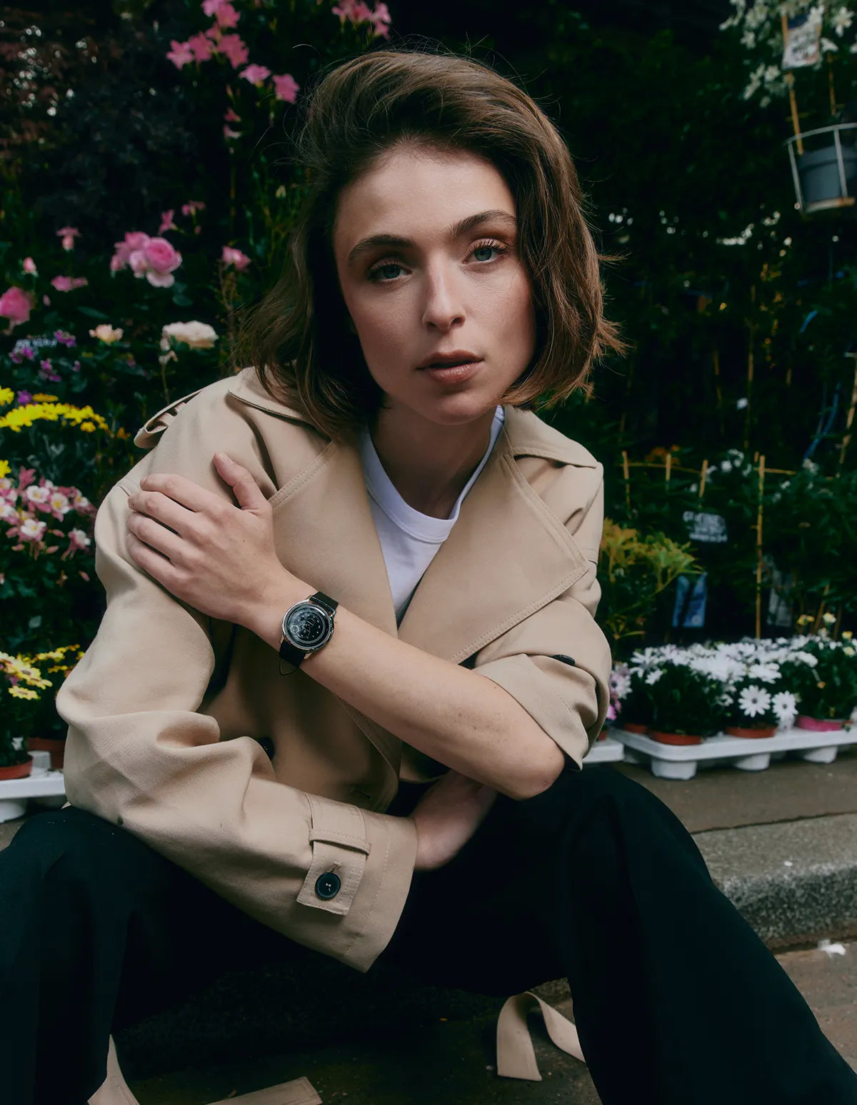Femme assise devant une boutique de fleurs portant un trench beige et une montre à cadran noir