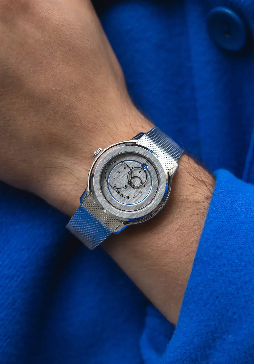 Montre Beableu Ecce Vesperal sur bracelet acier au poignet d'un homme avec une veste bleu 
